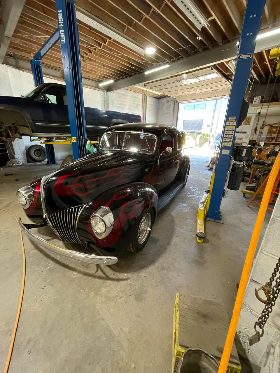 Black hot rod coupe with red flames on the shop floor between two lifts, a pickup up on a lift behind it